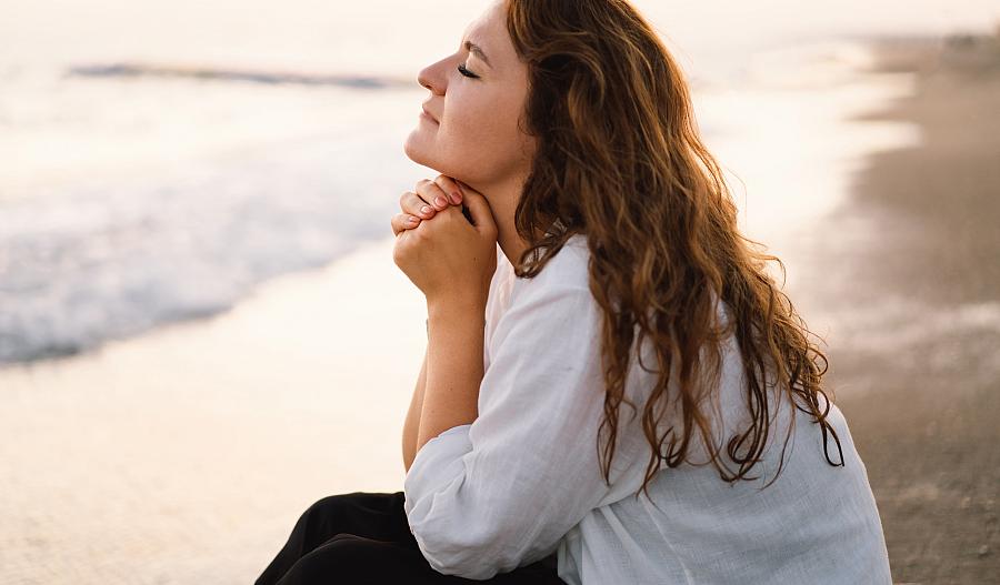 Imagen de una mujer con los ojos cerrados y las manos entrelazadas sentada junto al mar.