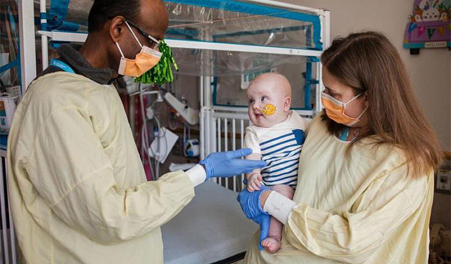 Una fotografía muestra a una doctora con un bebé en sus brazos, paciente de un tratamiento genético personalizado para tratar un trastorno potencialmente mortal. Otro doctor está saludando al bebé. Los doctores están usando mascarillas.