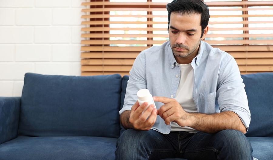 Una fotografía muestra a un hombre sentado en un sofá mientras lee la etiqueta de un frasco de medicamentos.